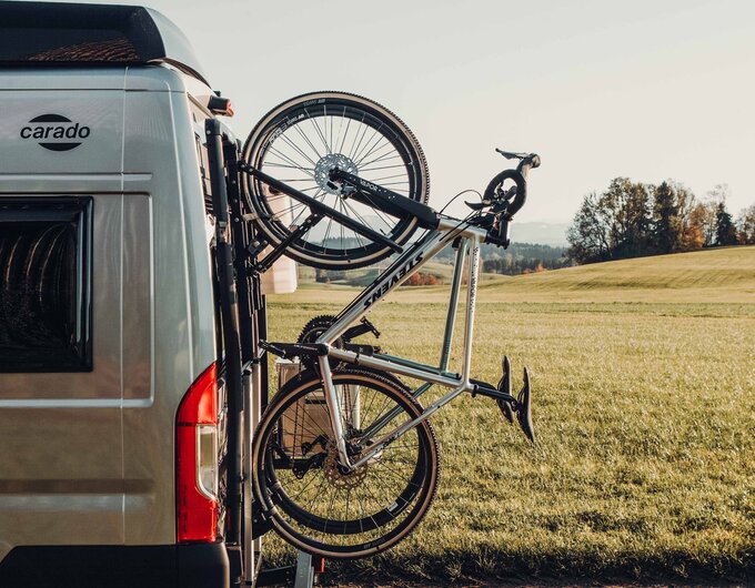 Rear of a Carado motorhome with a bike rack and a road bike parked at the roadside beside a meadow