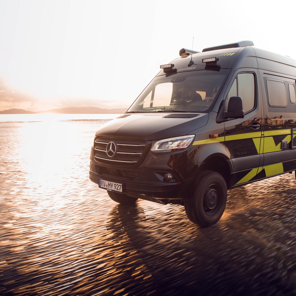 Mercedes Allrad-bobilen HYMER Grand Canyon S kjører raskt på en solfylt, våt strand. Omgivelsene er uklare på grunn av dynamikken.