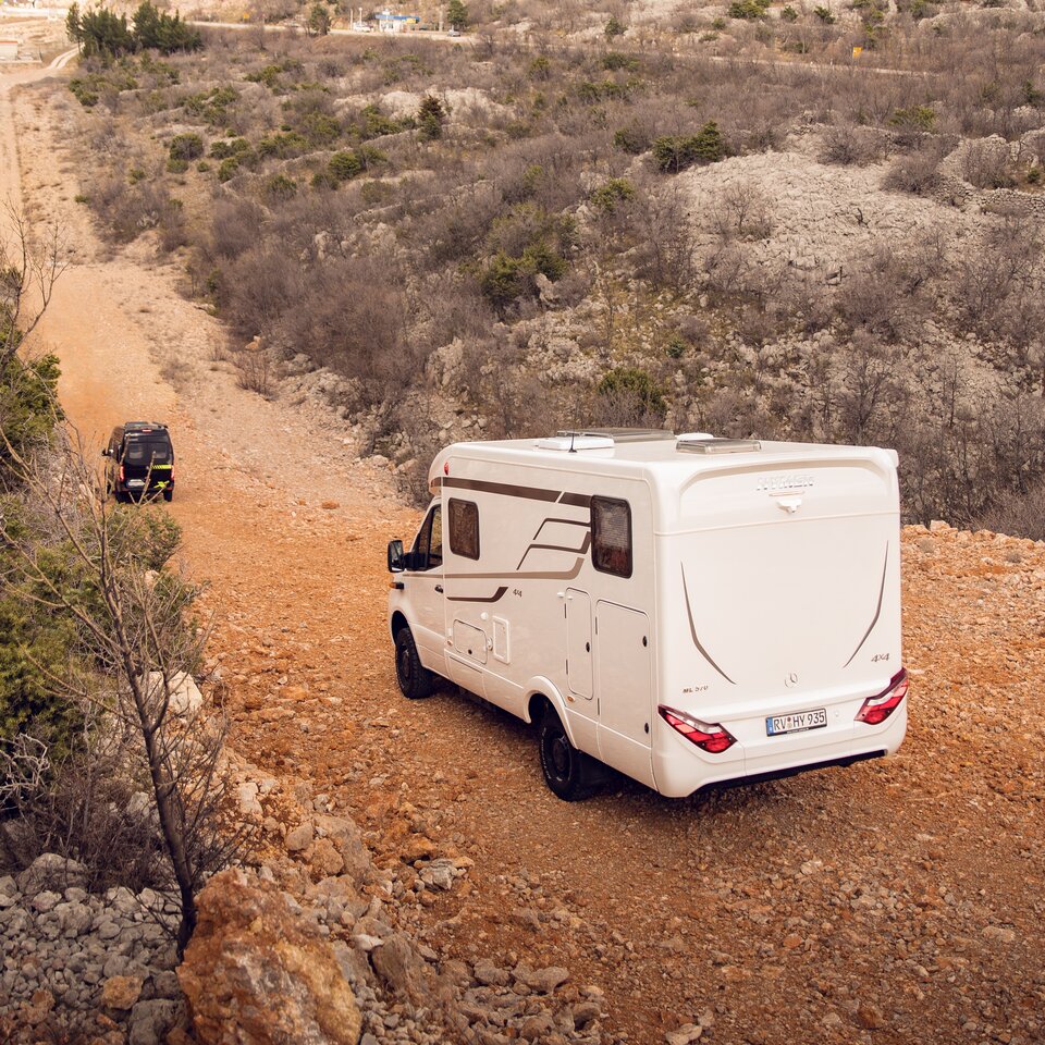 To 4x4 offroad-bobiler fra HYMER med Mercedes-førerkabin kjører på en kupert vei gjennom ujevn, steinete terreng med busker.