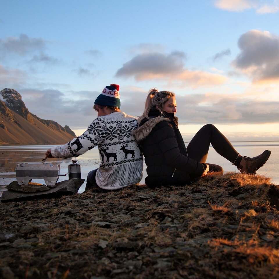 To personer i vinterklær sitter med en grill foran et vidstrakt landskap med fjell. Tatt på reise med bobil til Island.