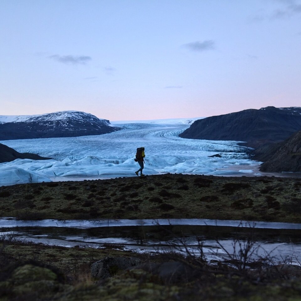 En person vandrer med ryggsekk gjennom landskap med gress, åser og snø. Bilde fra en reiseskildring til Island med ferge og bobil.