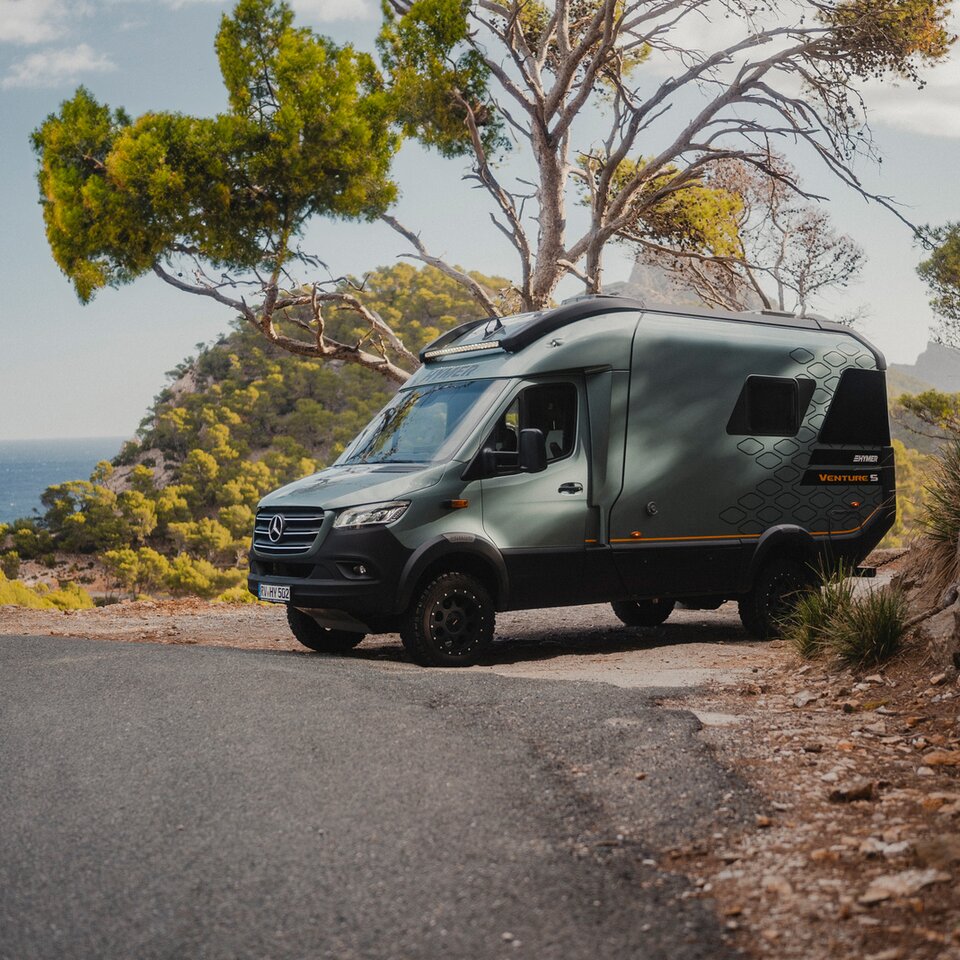 Vue de l'avant sur le Venture S, garé au bord de la route, entouré de montagnes, d'arbres et de la mer.
