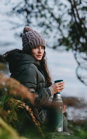 Een warm geklede vrouw met een thermosfles en een beker in haar hand zit op een rots in de natuur